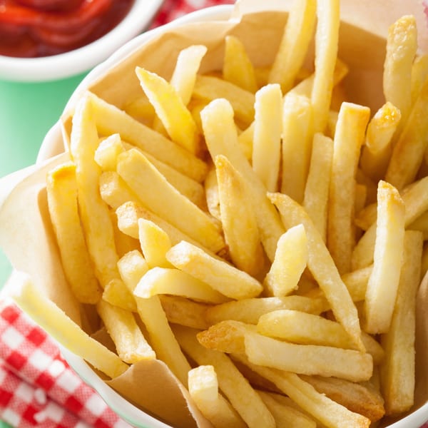 A close-up of golden french fries in a paper-lined cup, with ketchup in the background.
