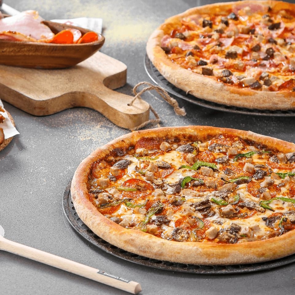 Two Milan's pizzas with various toppings, a wooden bowl of ingredients, and a pizza peel.